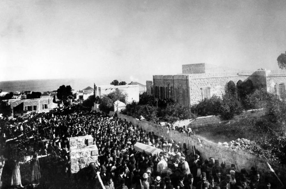 Ιστορική ασπρόμαυρη φωτογραφία με μεγάλο πλήθος ανδρών να ακολουθούν νεκρική πομπή, μεταφέροντας φέρετρο, σε έναν χωμάτινο δρόμο δίπλα σε πέτρινους τοίχους και κτίρια, με θέα τη θάλασσα στο βάθος.