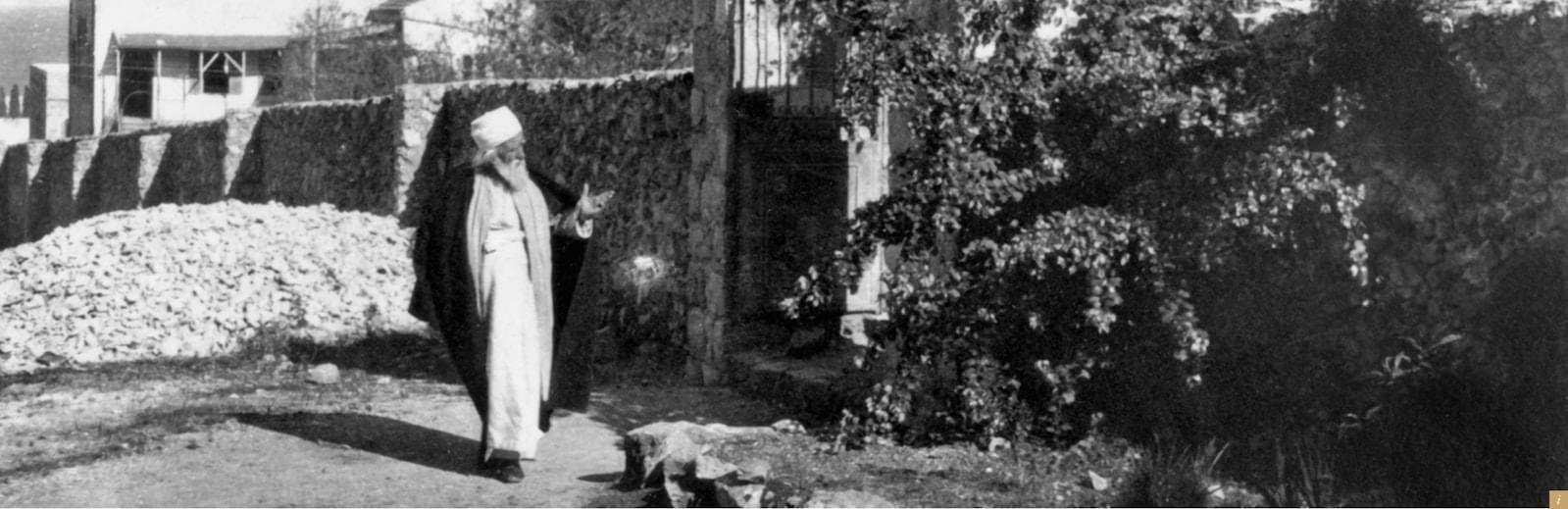 Ασπρόμαυρη ιστορική φωτογραφία του Αμπντούλ-Μπαχά, με λευκό χιτώνα και τάζι, όρθιου σε κήπο δίπλα σε πέτρινο τοίχο.