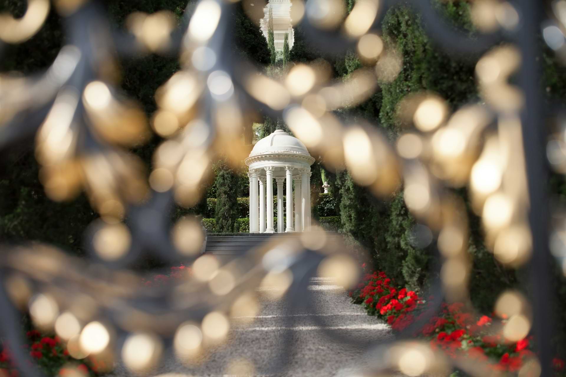 Λευκό κυκλικό μνημείο με κίονες και τρούλο στους Μπαχάι Κήπους, φωτογραφημένο μέσα από μια περίτεχνη σιδερένια πύλη.