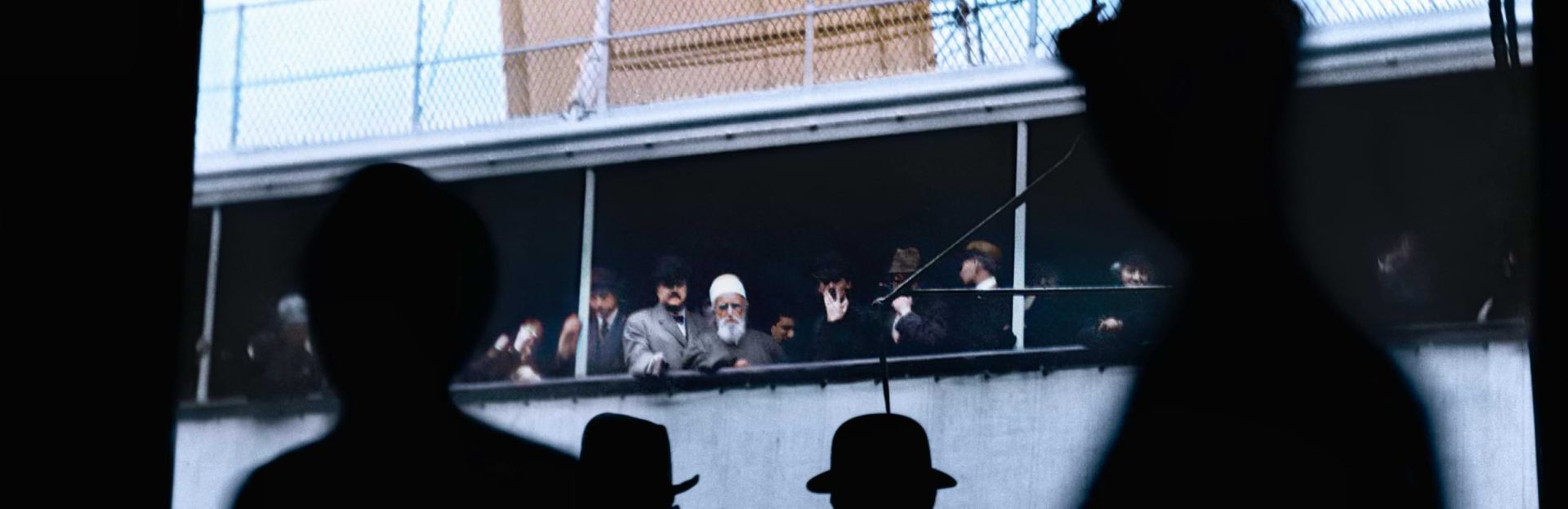 Έγχρωμη φωτογραφία του Αμπντούλ-Μπαχά σε κατάστρωμα πλοίου, ιδωμένη μέσα από τις σιλουέτες ανθρώπων στο προσκήνιο, με άλλους επιβάτες δίπλα του.