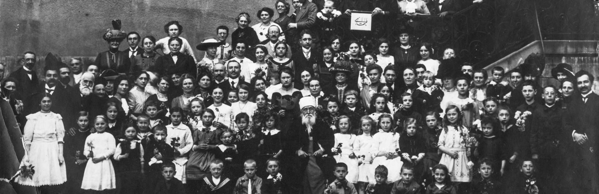 Ιστορική ασπρόμαυρη ομαδική φωτογραφία, που δείχνει ένα μεγάλο πλήθος ανδρών, γυναικών και παιδιών σε εξωτερικό χώρο με σκάλα, με τον Αμπντούλ-Μπαχά καθισμένο στο κέντρο