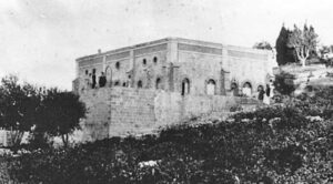 Ιστορική ασπρόμαυρη φωτογραφία ενός στιβαρού πέτρινου κτιρίου με τοξωτά παράθυρα και στρογγυλούς φεγγίτες, χτισμένου σε πλαγιά λόφου με βλάστηση