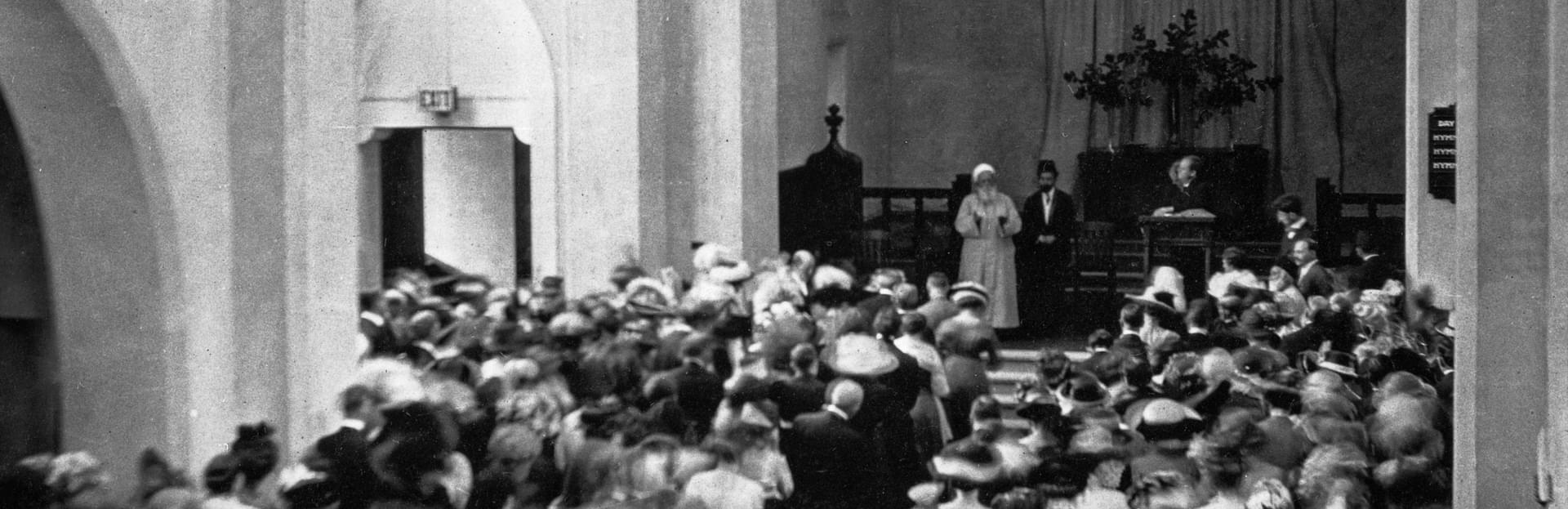 Ιστορική ασπρόμαυρη φωτογραφία του Αμπντούλ-Μπαχά να απευθύνεται σε ένα πολυπληθές εκκλησίασμα στο εσωτερικό ενός ναού με ψηλές αψίδες.